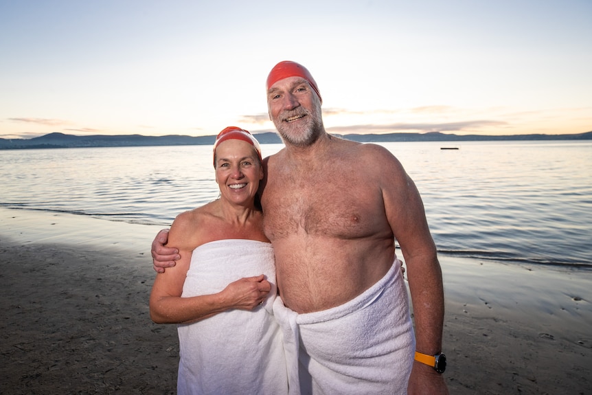 Two people stand on a beach wearing towels and red swimming caps.