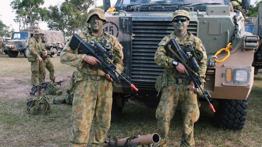 Two soldiers in camouflage uniform and face paint holding guns stand in front of an army tank