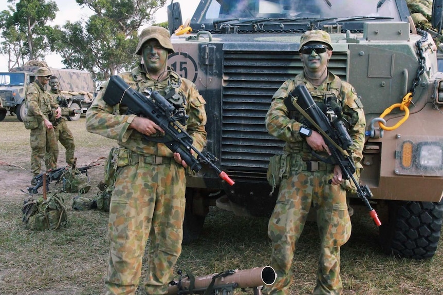 Two soldiers in camouflage uniform and face paint holding guns stand in front of an army tank