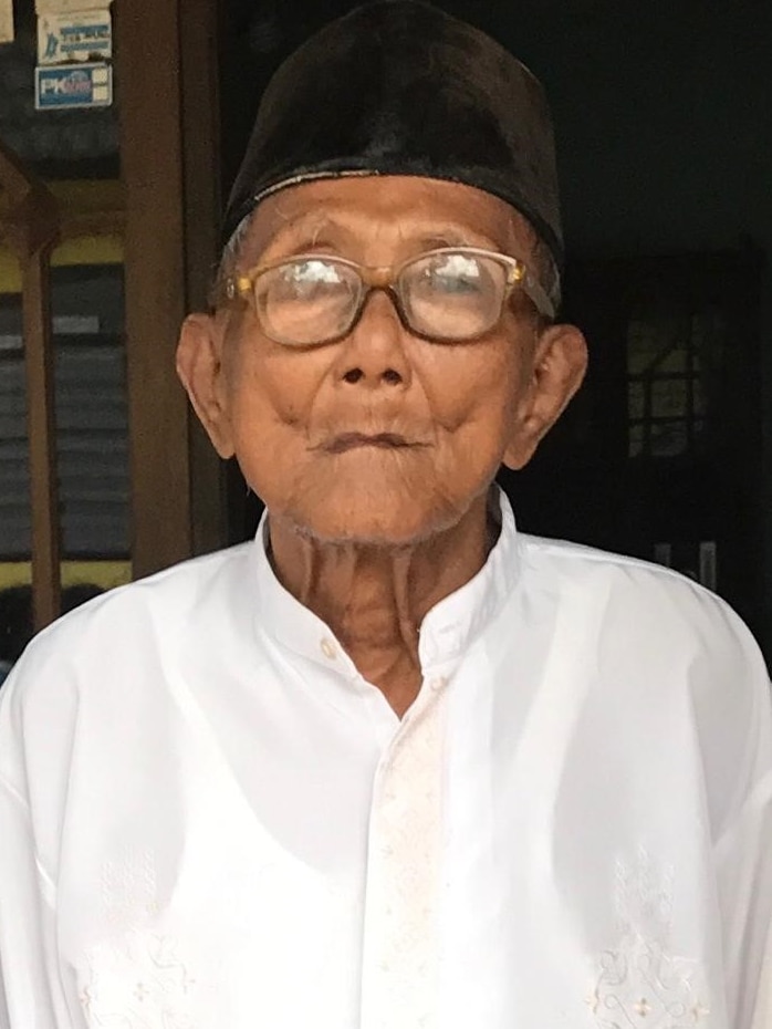 An elderly man wearing a white shirt, glasses and a black songkok cap.