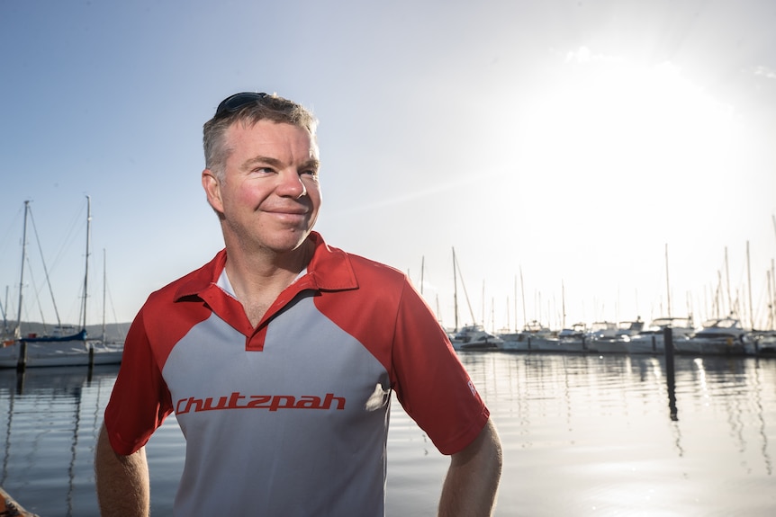 A man wearing a red and grey polo shirt stands at a sailing marina.