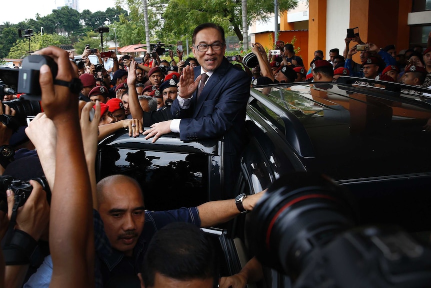 Anwar Ibrahim stands outside a car and waves as a crowd of photographers and police surround him.