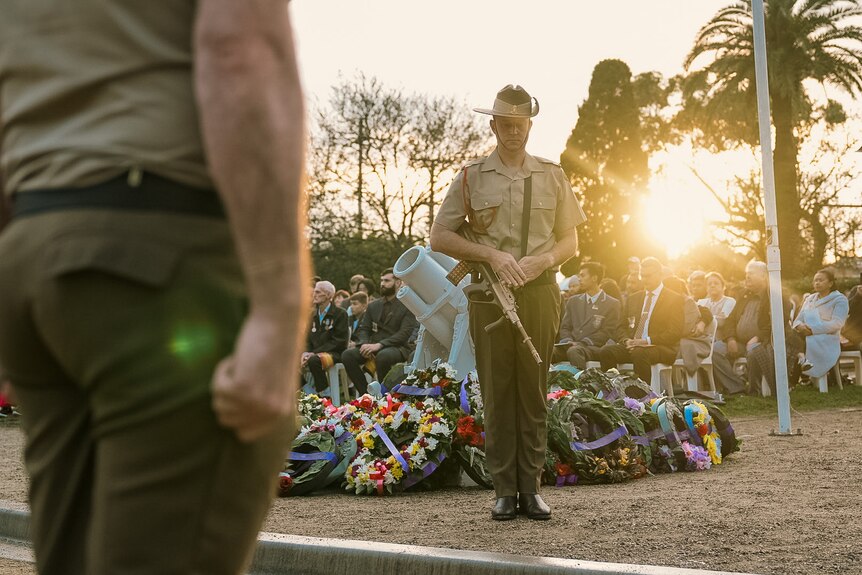 Two servicemen stand in front of a row of wreaths and a crowd gathered to mark Anzac Day.