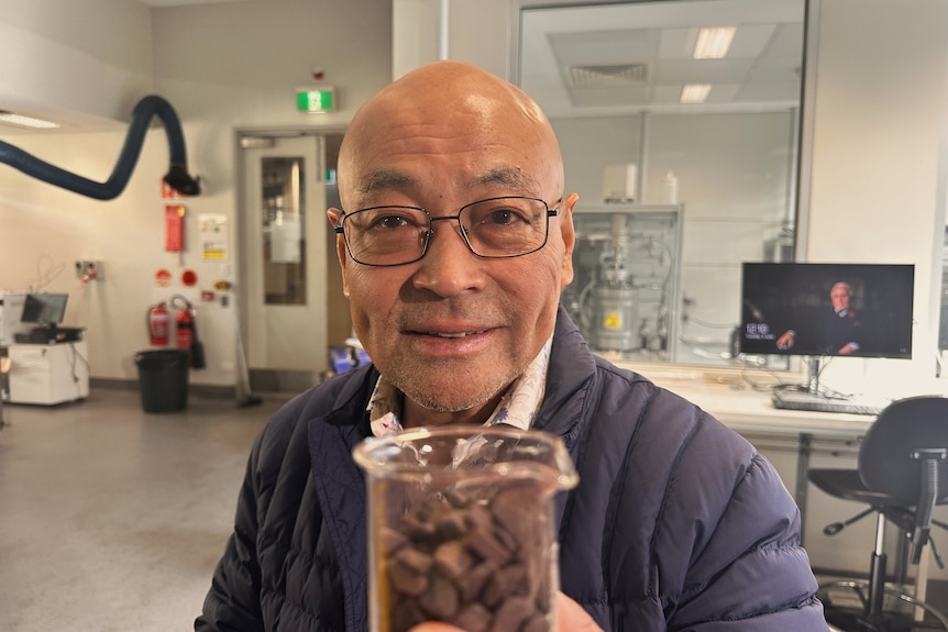 A professor named Dongke Zhang holds a beaker full of iron ore towards the camera while smiling slightly. 