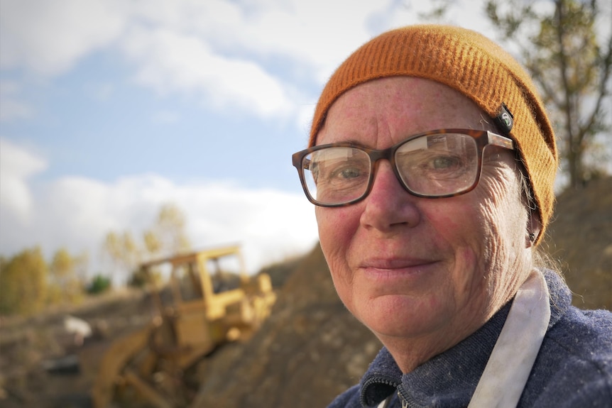 Close shot of a woman wearing a beanie smiling to camera, earth moving machinery a blur in the background.