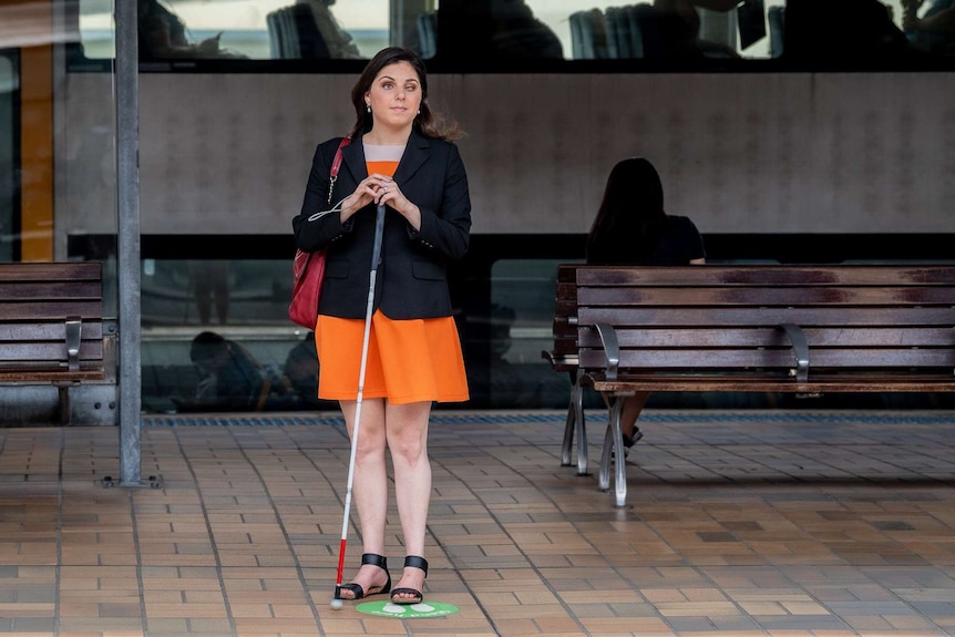 Nas stands on a train platform with her cane.