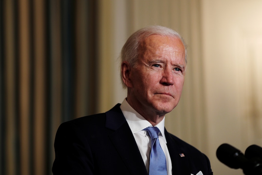 President Biden stands in front of a microphone wearing a black suit and blue tie with an American flag pinned to his lapel