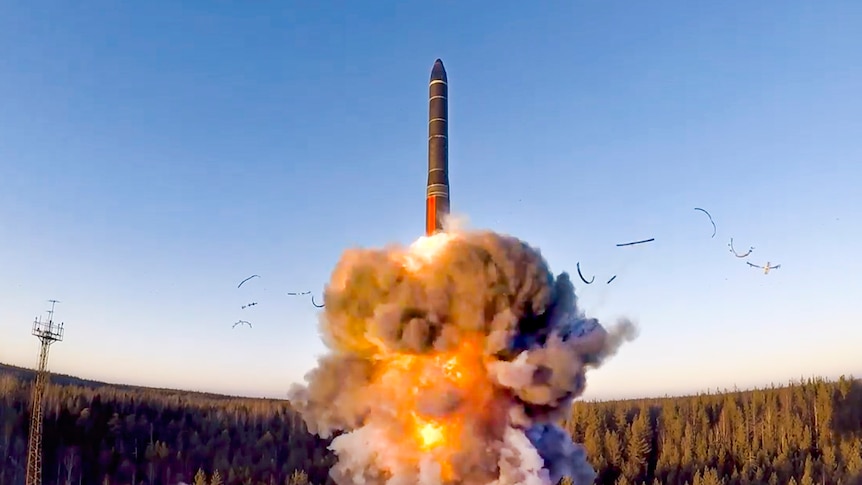 A missile is launched into the blue sky. Plumes of grey and orange smoke are underneath it. 