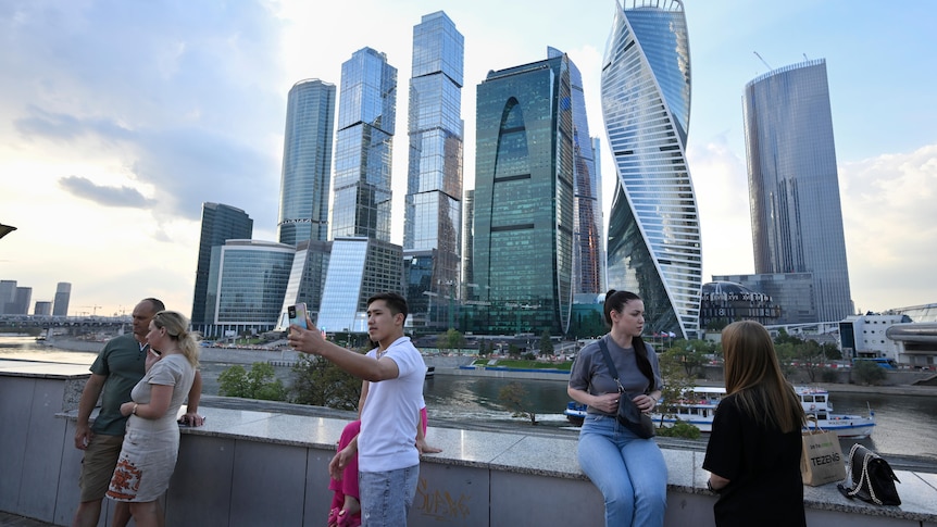 People stand and take photos at embankment of the Moscow River with the "Moscow City" business district in the background.