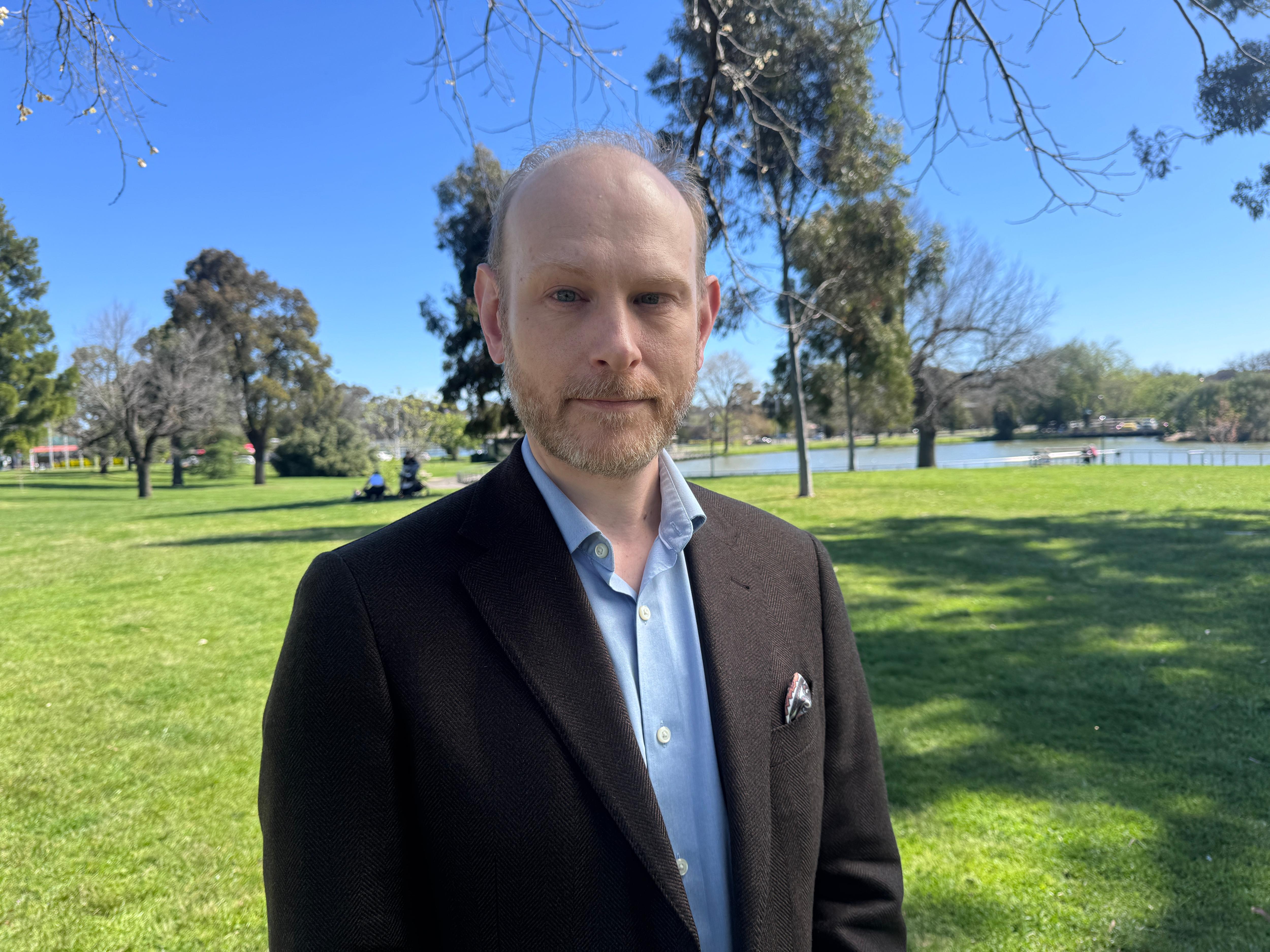 A man in a suit and blue button up shirt and beard, stands in a green park.