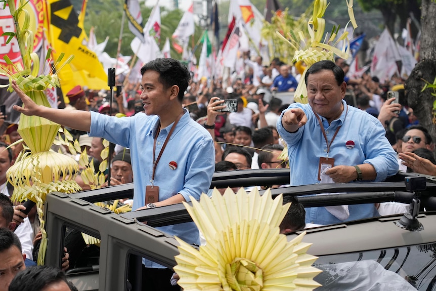 Prabowo Subianto and a young man stand in an open top car waving and pointing at a crowd of supporters