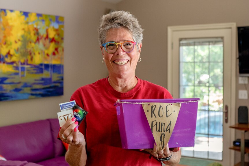 A middle-aged woman with rainbow-rimmed glasses smiles while clutching condoms and a bucket labelled ROE FUND