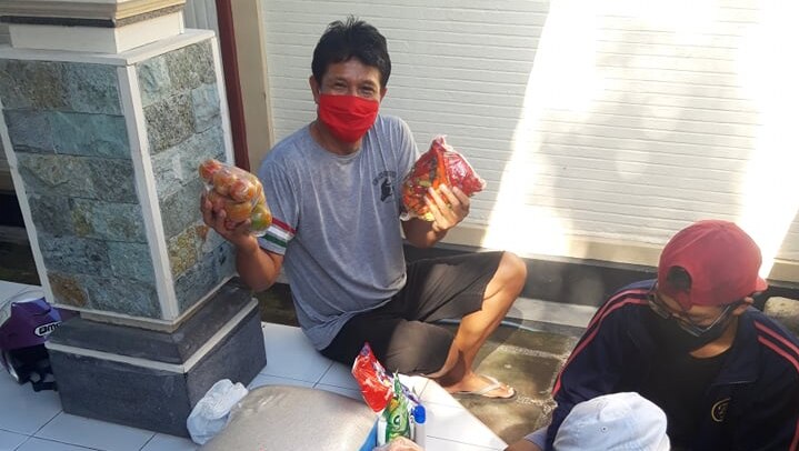 A man wearing a mask holding up food he has just received, with other necessities like rice and oil by his feet.
