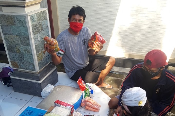A man wearing a mask holding up food he has just received, with other necessities like rice and oil by his feet.