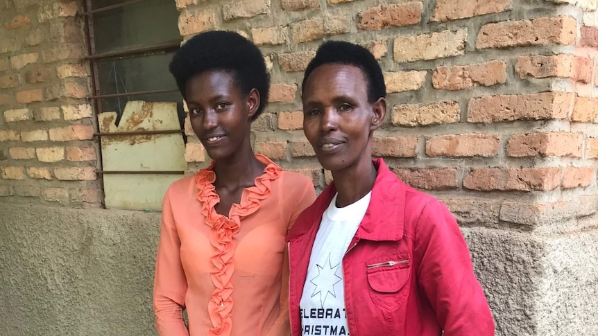 A young woman stands next to her mother against a wall. They are both smiling.