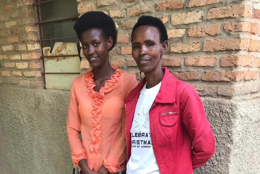 A young woman stands next to her mother against a wall. They are both smiling.