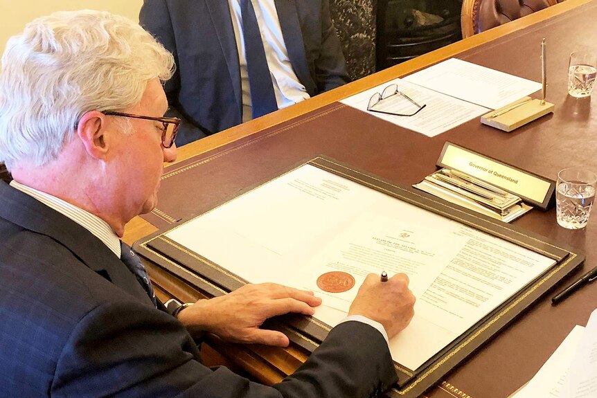 Queensland Governor Paul de Jersey sitting at a table and signing a document.