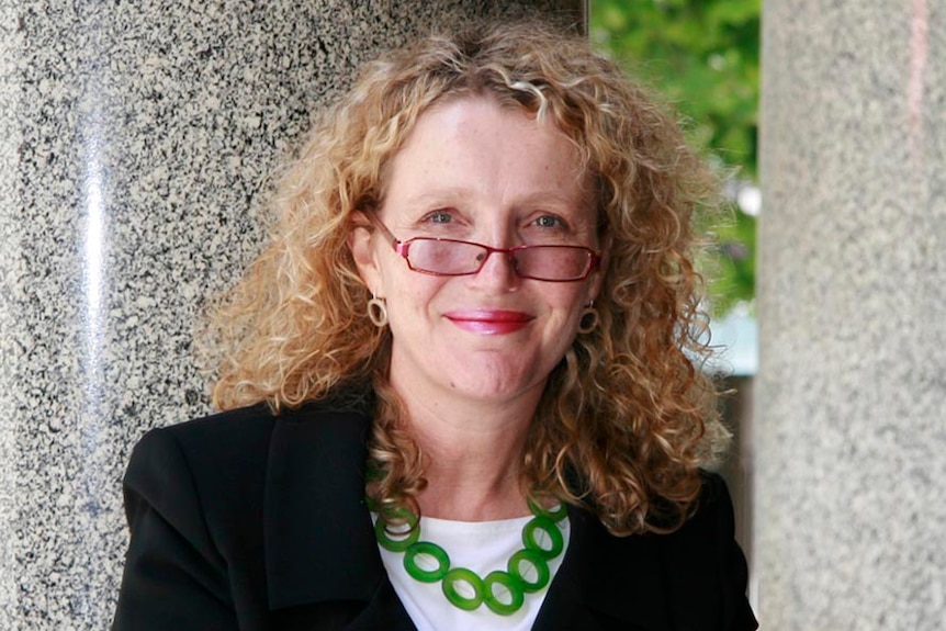 A woman with frizzy, ginger hair and glasses, wearing a dark blazer.
