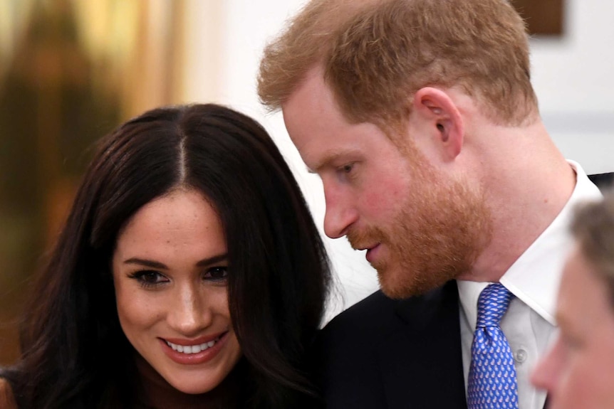 Prince Harry turns to Meghan Markle, speaking in her ear, as she smiles.