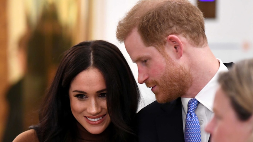 Prince Harry turns to Meghan Markle, speaking in her ear, as she smiles.