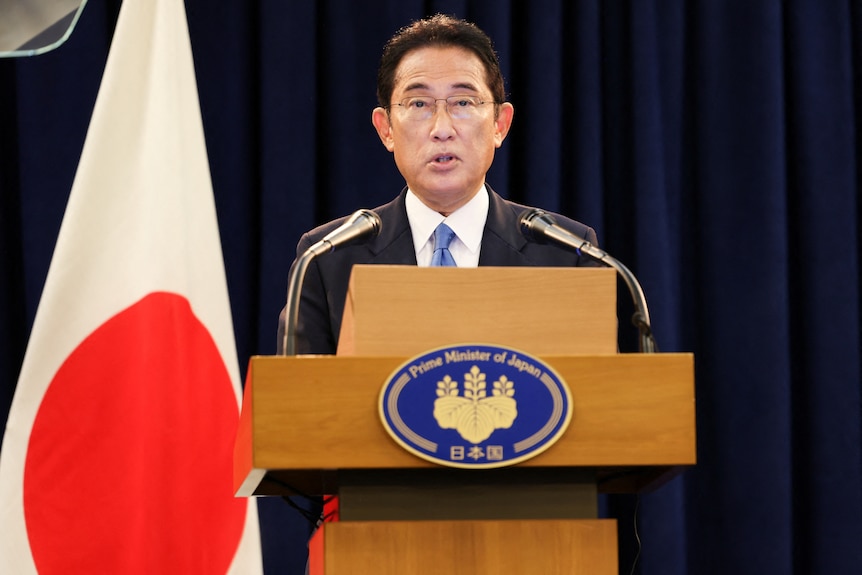 Fumio Kishida speaking at a lectern next to a Japanese flag with a blue curtain behind him
