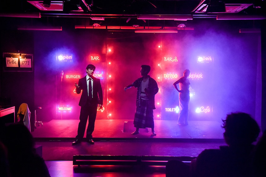 A man wears black tie and suit gestures near two silhouetted figures backlit by magenta and purple neon sigs on hazey stage.