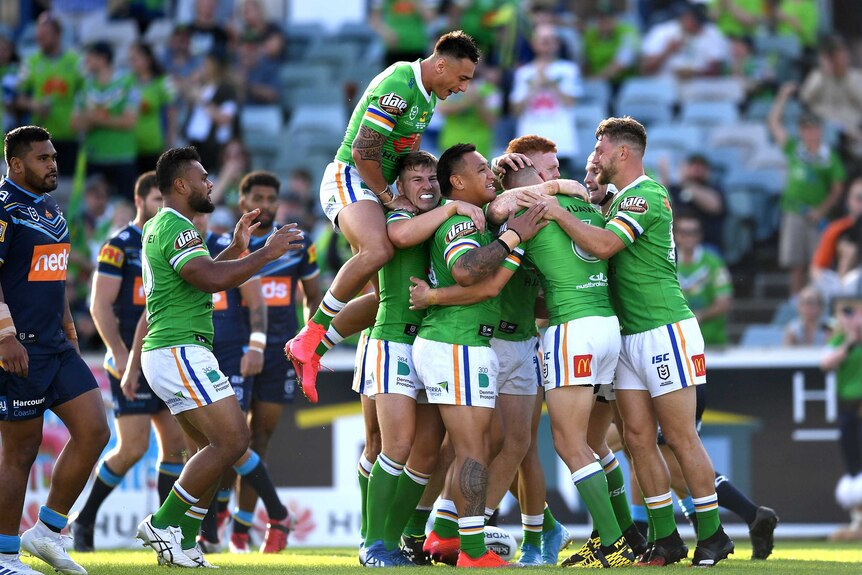 Canberra Raiders players in green jerseys celebrate a try in a pack as one player jumps atop the group.