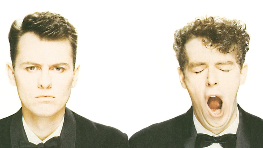 Two members of Pet Shop Boys wear tuxedos and stand before a white background. One of them yawns.