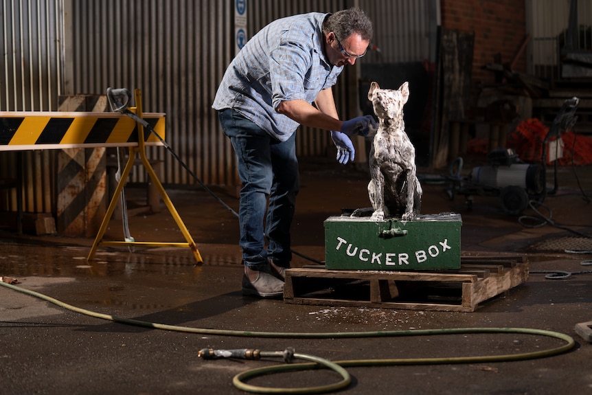 A man with gloves works on a statue of a dog sitting on a green tuckerbox.