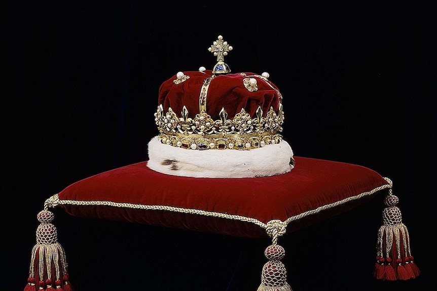 A golden crown with a red velvet cap and a white fur trim sits on a red cushion. 