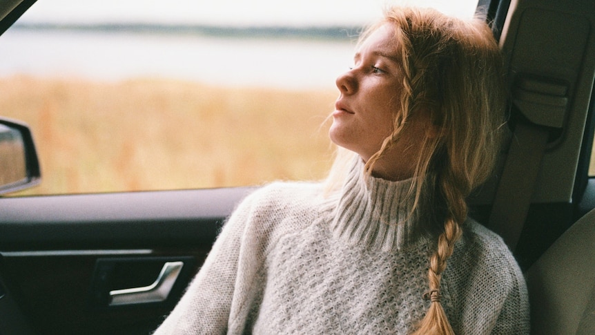 A young blonde woman stares into space in her car. She is wearing a oatmeal coloured knit.