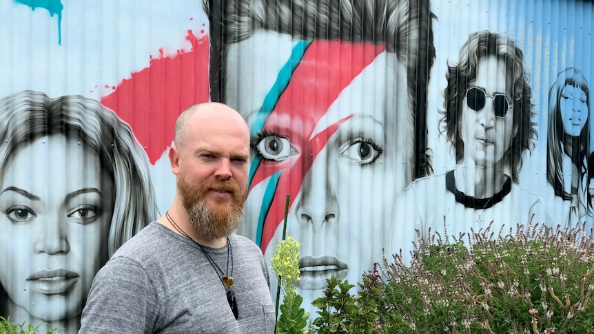 Man standing in front of a very large mural, painted on corrugated iron which features portraits of musicians.