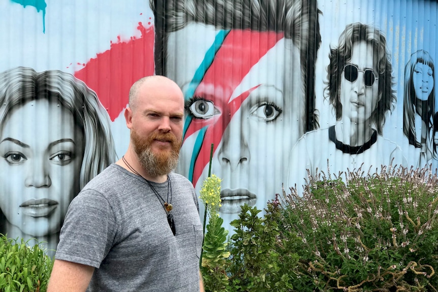 Man standing in front of a very large mural, painted on corrugated iron which features portraits of musicians.
