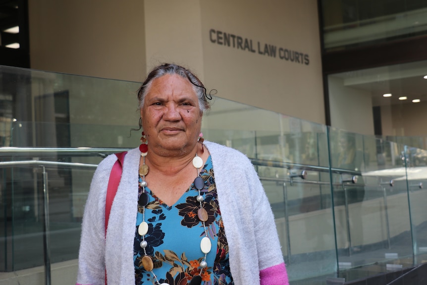 Eine ältere indigene Frau, die ein blaues Blumenoberteil und eine graue Strickjacke trägt, posiert für ein Foto vor dem Gericht in Perth.