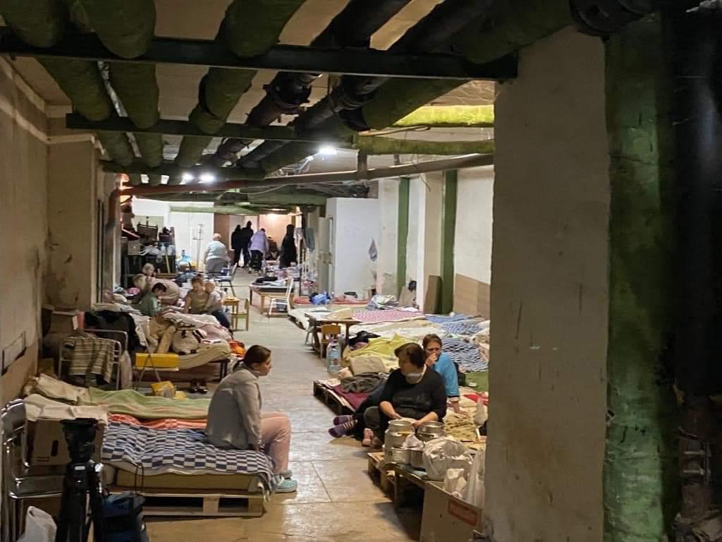 People sit in a bunker in Ukraine in a sparse covered room with stretches and a few items around