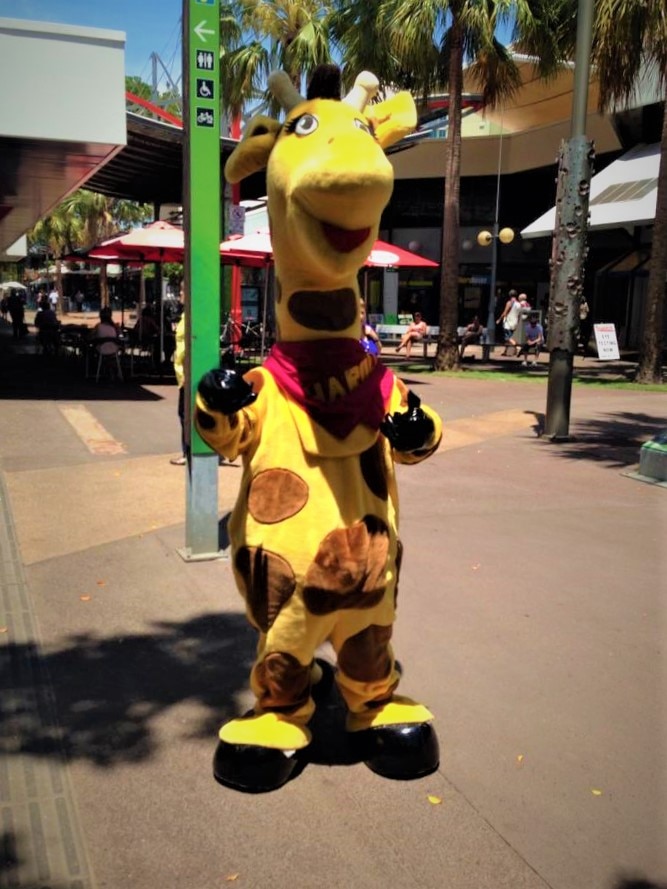 A giraffe mascot giving the thumbs up on a pedestrian street.