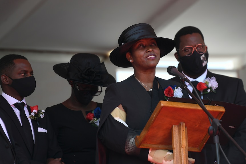 A middle-aged Haitian woman wearing a black dress and hat stands behind a lectern as she speaks into a microphone.