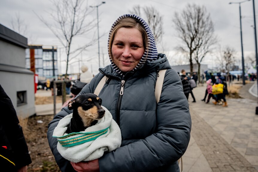 a woman in a grey jumper and hoodie holds a small dog in a blanket in her arms as refugees walk behind in Lviv