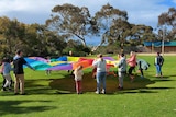 A group of children in a large circle play with a rainbow-coloured parachute on the grass