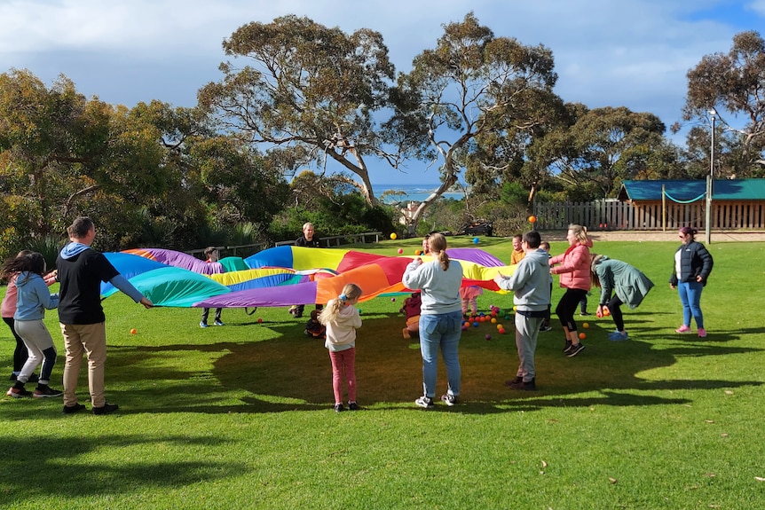 A group of children in a large circle play with a rainbow-coloured parachute on the grass