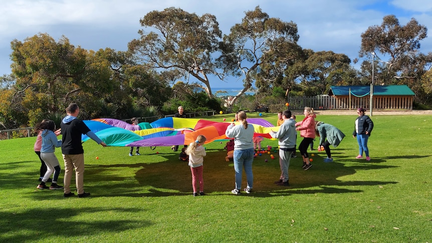 A group of children in a large circle play with a rainbow-coloured parachute on the grass