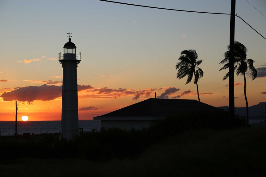 The sun setting over the ocean creates a silhouette of a lighthouse and palm tree