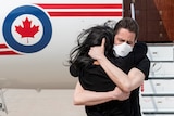 A man in a mask hugs his wife in front of a plane.