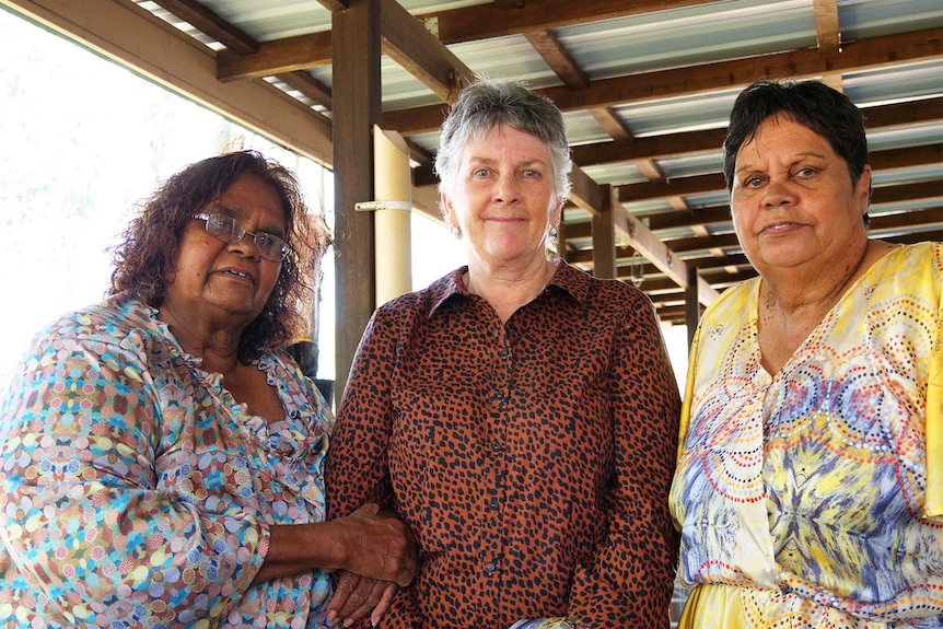 Two older Aboriginal women in colourful silk shirts with dot designs on them stand with another lady on a verandah.