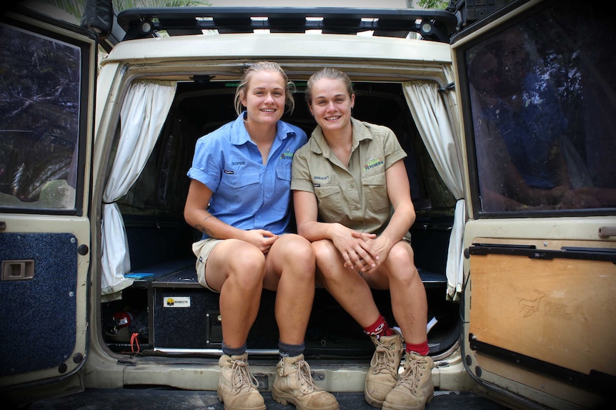 Two 23-year-old blonde twins sit on the back of a truck, one in a blue shirt, the other in a khaki shirt.