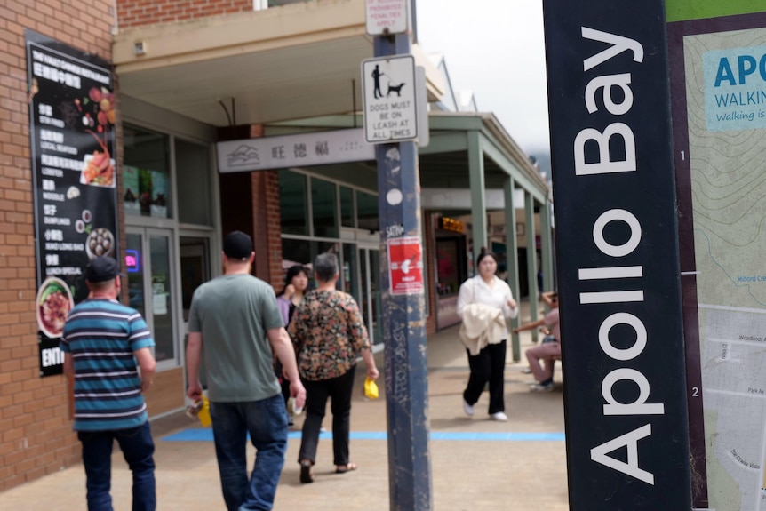 People walk along a footpath next to a sign that says 'Apollo Bay'.