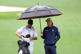 Two men stand underneath an umbrella on a golf course as it rains