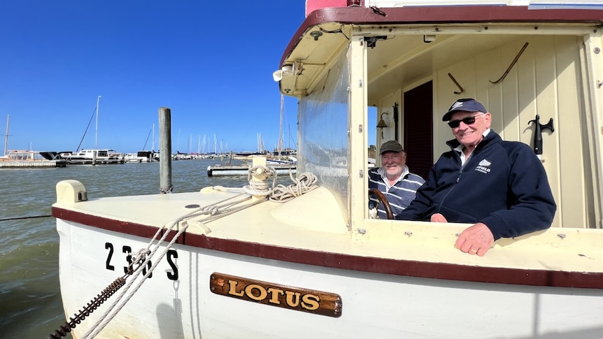 Two men seated near the wheel of a cream and burgundy-coloured restored wooden boat with the name 'Lotus' on the hull