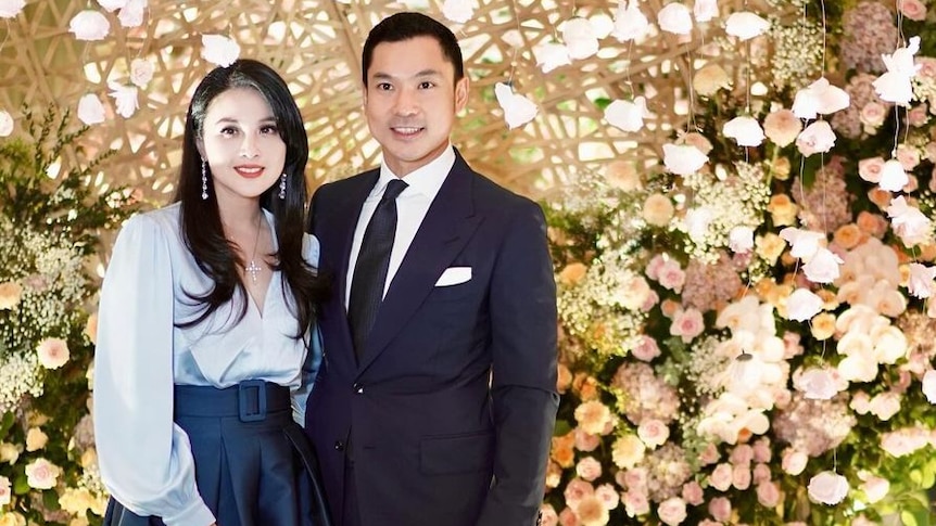 Sandra Dewi smiles in a blue blouse and skirt with a man wearing a dark suit and black tie in front of flowers.
