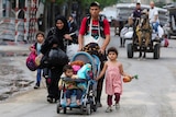 A man, woman and three young children walk down a street carrying bags and backpacks, youngest child sits in a pram